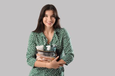 Gri arka planda makyaj fırçaları olan kozmetik ürünler ve kozmetik poşetler taşıyan güzel genç bir kadın.
