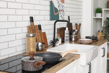 Mutfaktaki beyaz tuğla duvarın yanındaki tezgahta soba, lavabo ve çatal bıçak var.