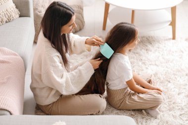 Genç anne evde yerde otururken kızının saçlarını tarıyor.