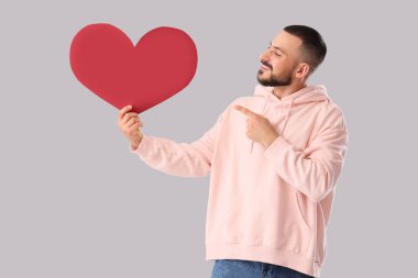 Yakışıklı genç adam gri arka planda kağıt kalbe işaret ediyor.
