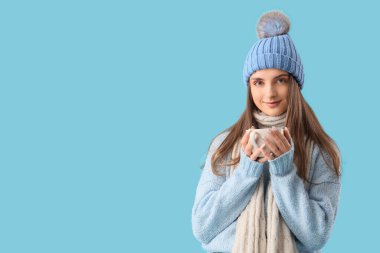 Güzel, genç bir kadın, şık ve sıcak giysiler içinde. Mavi arka planda bir fincan sıcak çay.