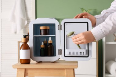 Masadaki küçük kozmetik buzdolabından yeşil arka planda yüz kaldıran kadın elleri. Yakın çekim.