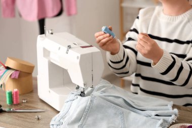 Genç bayan terzi atölyenin yanındaki masanın yanında kot şort dikmek için dikiş makinesi kullanıyor.
