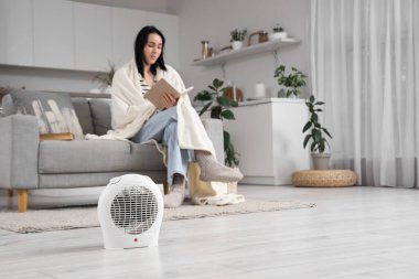 Mutfakta kitap okuyan bir kadına karşı yerde elektrikli vantilatör var.