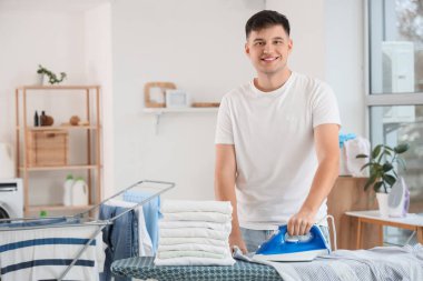 Yakışıklı genç adam evde giysileri ütülüyor.