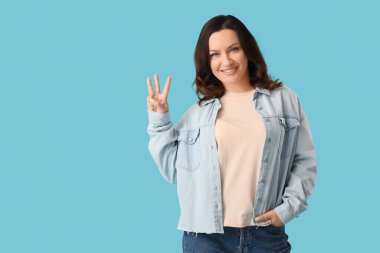 Güzel, olgun, mutlu bir kadın mavi arka planda üç parmağını gösteriyor.