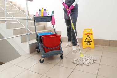 Araba dolusu temizlik malzemesi taşıyan kadın hademe merdivenleri paspaslıyor.