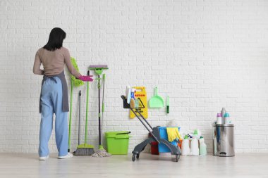 Beyaz tuğla duvarın yanında temizlik malzemesi ve uyarı levhası taşıyan kadın hademe.