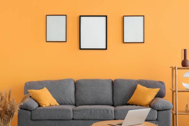 Koltuk, dizüstü bilgisayarlı masa ve sarı duvarda boş posterler.