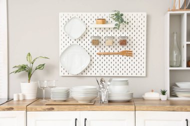 Mutfaktaki beyaz duvarın yanındaki tezgahlarda ev bitkisi, tabak, tabak ve çatal bıçak takımı var.