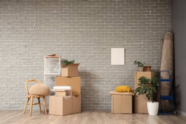 İçinde ev bitkileri olan karton kutular, rulo halı ve taşınma gününde oturma odasındaki gri tuğla duvarın yanında hasır sandalye.