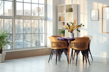 Yemek odasının içinde, pencerenin yanındaki masada güzel çiçekler ve şık mor kumaşlar var.