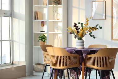 Yemek odasının içinde, pencerenin yanındaki masada güzel çiçekler ve şık mor kumaşlar var.