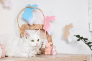 Masada oyuncak tavşanlarla oturan sevimli beyaz kedi ve evde tavşan şeklinde kağıt çelenkler. Paskalya kutlaması