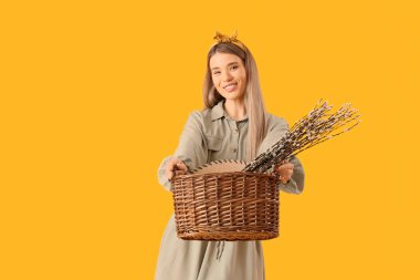 Beautiful young woman holding wicker basket with pussy willow branches on yellow background