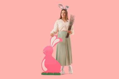 Beautiful young woman in bunny ears with pussy willow branches and paper rabbit on pink background