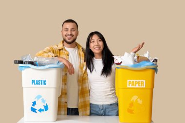 Bej arka planda çöp bidonlarına yerleştirilmiş genç bir çift. Küresel Geri Dönüşüm Günü