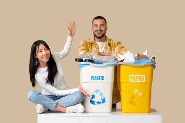 Bej arka planda çöp bidonlarına yerleştirilmiş genç bir çift. Küresel Geri Dönüşüm Günü