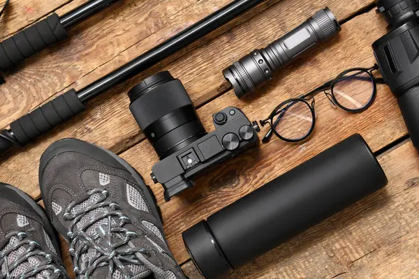 Modern fotoğraf makinesi, termos, el feneri, ayakkabılar ve tahta arka planda yürüme direkleriyle kompozisyon, yakın plan.