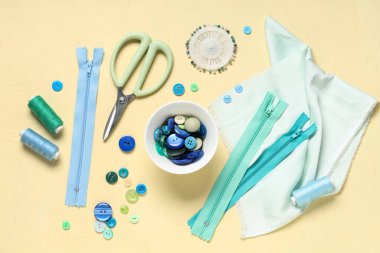 Renkli düğmeler, kumaş örnekleri, iplik makaraları, makas ve fermuarlar sarı arkaplanda.