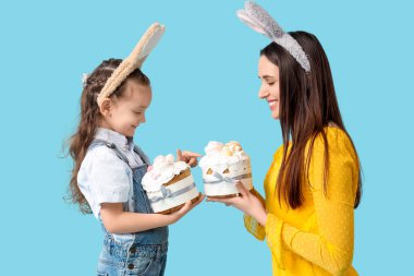 Güzel genç bir kadın ve küçük tatlı kızı tavşan kulaklı ve mavi arka planda Paskalya pastaları var.