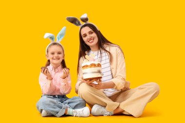 Tavşan kulaklı güzel genç bir kadın. Elinde Paskalya pastası ve sarı arka planda çikolata yumurtaları olan tatlı küçük kızı var.