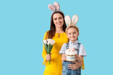 Bir buket lale çiçeğiyle ve mavi arka planda Paskalya pastası tutan küçük tatlı kızıyla tavşan kulaklı güzel bir kadın.