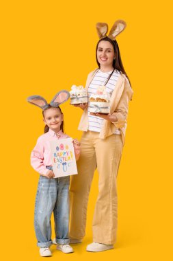 Paskalya pastası ve sarı arka planda tebrik kartı tutan sevimli küçük kızı ile tavşan kulaklı güzel bir genç kadın.