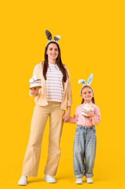 Güzel genç bir kadın ve sarı arka planda Paskalya pastaları olan tavşan kulaklı küçük tatlı kızı.