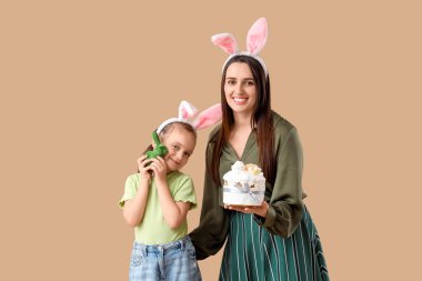Paskalya pastası ve kahverengi arka planda oyuncak tavşan tutan sevimli küçük kızı ile tavşan kulaklı güzel bir genç kadın.