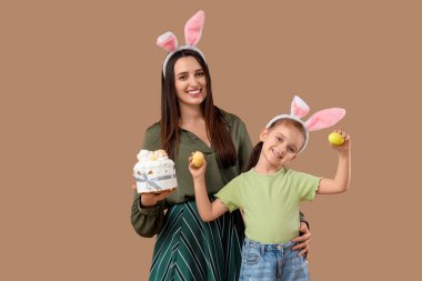 Paskalya pastası ve kahverengi arka planda boyalı yumurta tutan sevimli küçük kızı ile tavşan kulaklı güzel bir genç kadın.