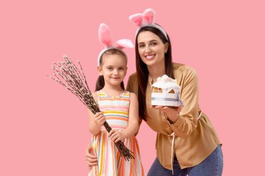 Beautiful young woman in bunny ears with Easter cake and her cute little daughter holding pussy willow branches on pink background