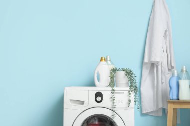 Çamaşır deterjanı şişeleriyle modern çamaşır makinesi ve mavi duvarın yanında ev bitkisi.