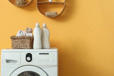 Turuncu duvarın yanında deterjan şişeleri ve hasır sepeti olan modern çamaşır makinesi.