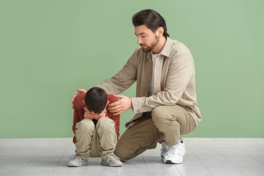 Genç baba, yeşil arka planda tartıştıktan sonra üzgün oğlunu sakinleştirmeye çalışıyor.