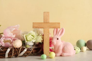 Tahta haç, boyalı yumurta, tavşan, çiçekler ve çelenk beyaz masada, sarı grunge arka planda. Paskalya kutlaması