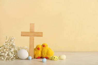 Piliçlerle ahşap haç, beyaz masaya boyanmış yumurta ve çiçekler, sarı grunge arka plana karşı. Paskalya kutlaması