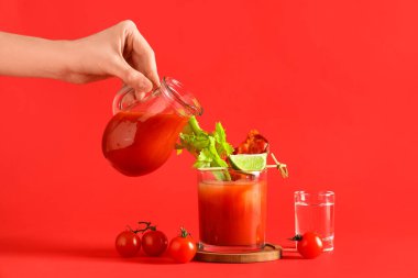 Kadın kırmızı arka planda domuz pastırması ve kereviz ile sürahiden bir bardak Bloody Mary kokteyline domates suyu döküyor.