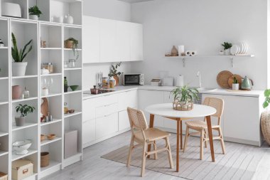 Beyaz tezgahlı, masalı, ev bitkilerli ve raf üniteli şık bir mutfak.