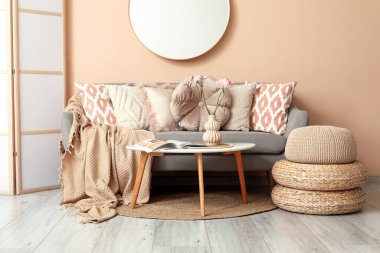 Beyaz perdeli, kahve masalı ve bej duvarın yanındaki gri koltukta pembe yastıklı şık bir oda.