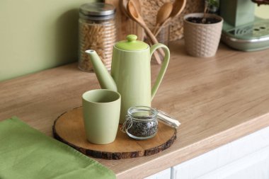 Mutfaktaki yeşil duvarın yanındaki masada bir kavanoz kuru çay ve fincanla demlik.