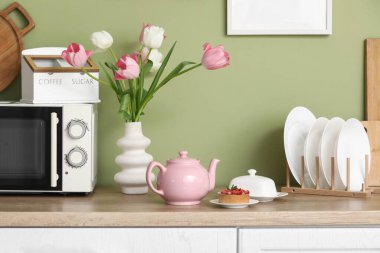 Pembe çaydanlık, lezzetli bir tatlı ve güzel lale çiçekleri mutfaktaki yeşil duvarın yanındaki masada.