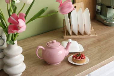 Pembe çaydanlık, lezzetli bir tatlı ve güzel lale çiçekleri mutfaktaki yeşil duvarın yanındaki masada.