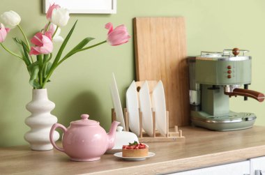 Pembe çaydanlık, lezzetli bir tatlı ve güzel lale çiçekleri mutfaktaki yeşil duvarın yanındaki masada.