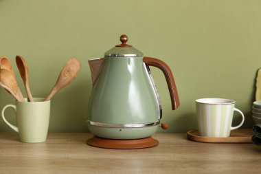 Mutfaktaki yeşil duvarın yanındaki masada fincanlı elektrikli çaydanlık ve çatal bıçak.