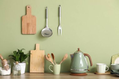 Mutfaktaki yeşil duvarın yanındaki masada fincanlı elektrikli çaydanlık ve çatal bıçak.