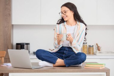 Bir fincan kahve ve dizüstü bilgisayarlı genç kız mutfakta online olarak çalışıyor.