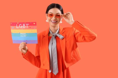 Elinde gökkuşağı bayrağı olan güzel bir genç kadın turuncu arka planda LGBT Övünme Ayı