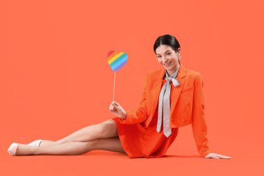 Turuncu arka planda oturan gökkuşağı kalpli güzel bir genç kadın. LGBT kavramı