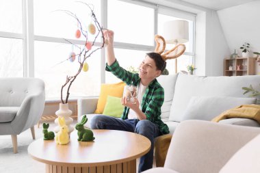 Tavşan kulaklı genç çocuk evde paskalya yumurtalarıyla ağaç dallarını süslüyor.